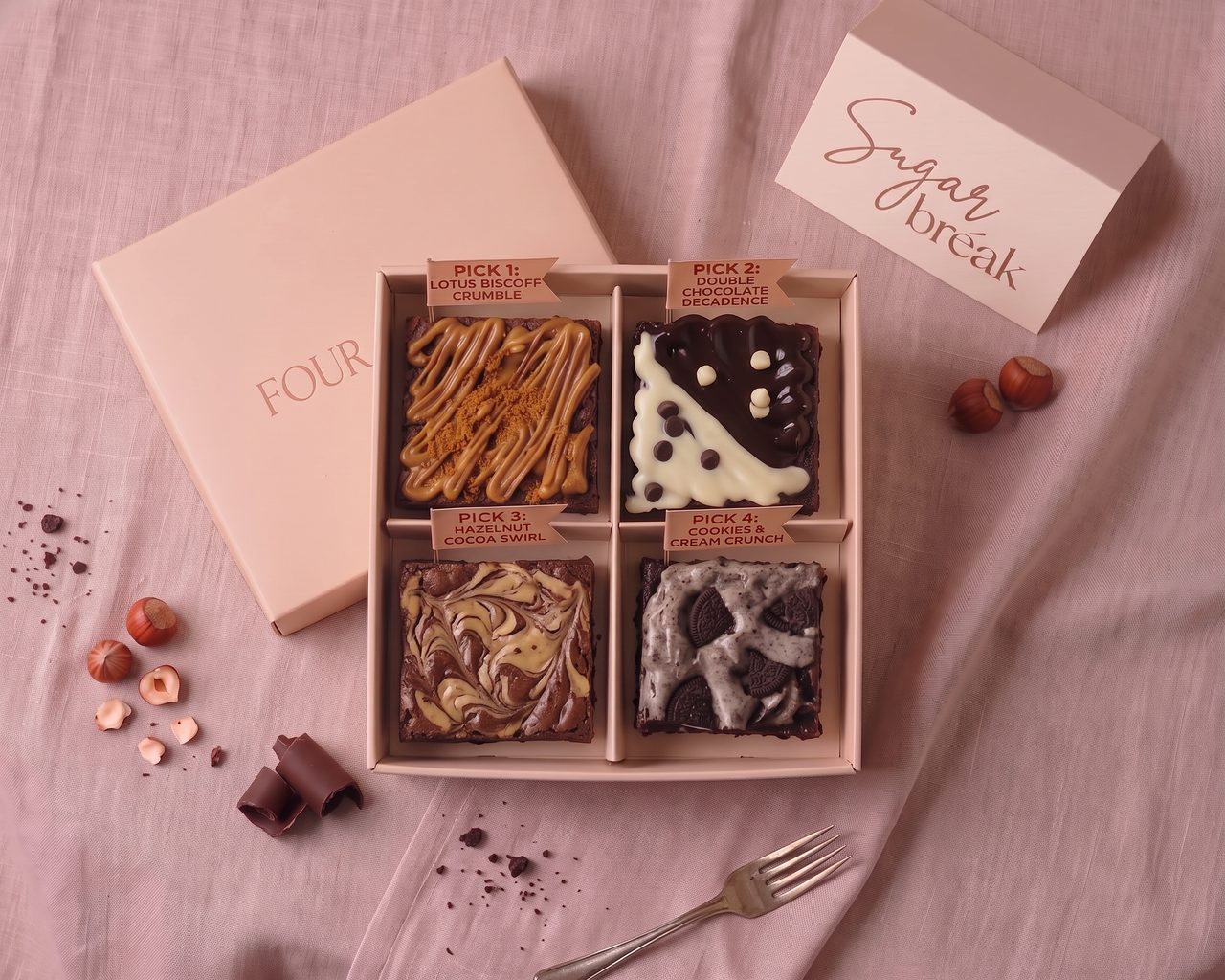 Four Galore from above, an open pink box with four brownies (Lotus Biscoff Crumble, Double Chocolate Decadence, Hazelnut Cocoa Swirl, Cookies & Cream Crunch), each with a pick flag, beside its lid and a Sugar bréak card
